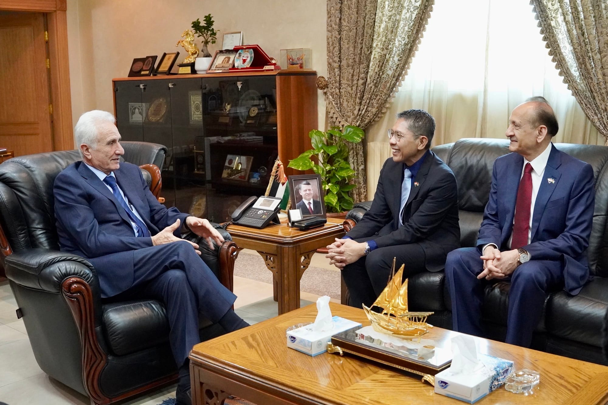 Three men in suits sit in an office setting, two on a leather sofa. Photos and decorations are on the tables.